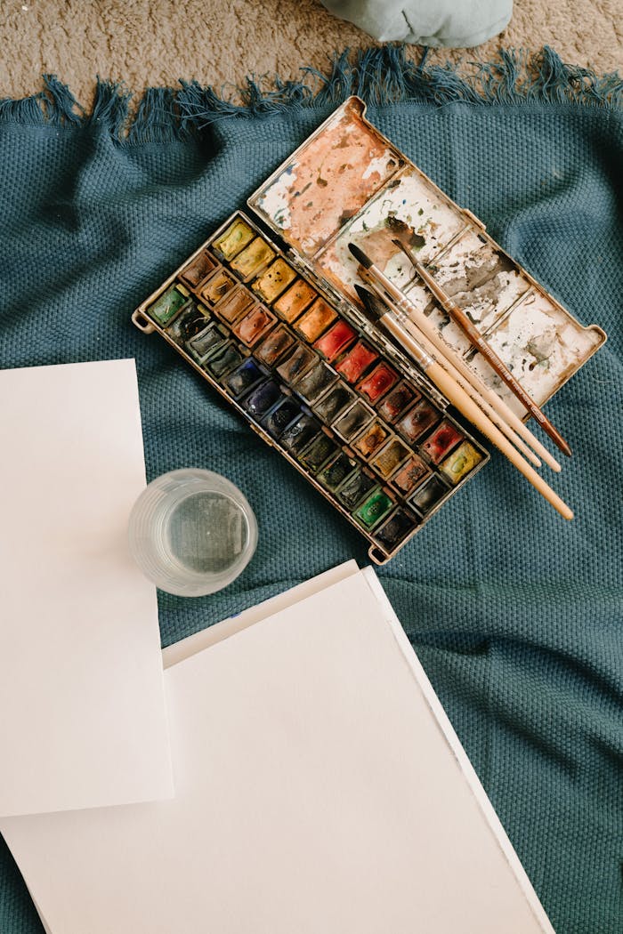 Flat lay of watercolor paints, brushes, and blank paper on a fabric background, ideal for creative inspiration.