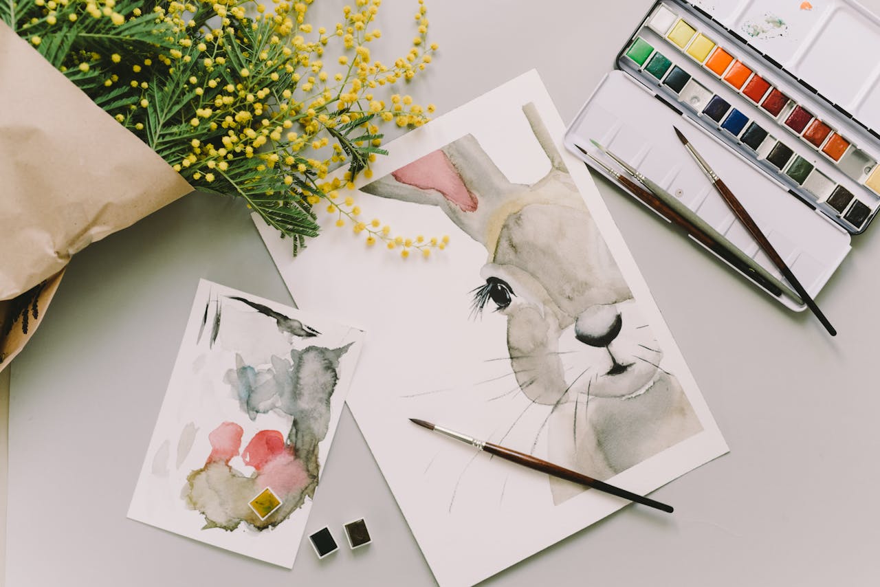 A creative flatlay featuring watercolor paintings of an Easter bunny with mimosa flowers and art supplies.