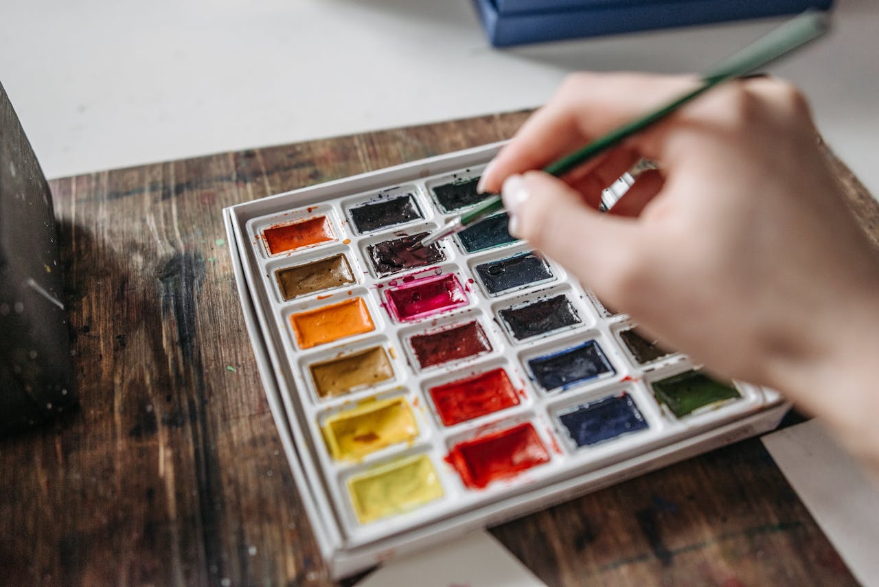 An artist's hand selecting a watercolor from a vibrant palette.